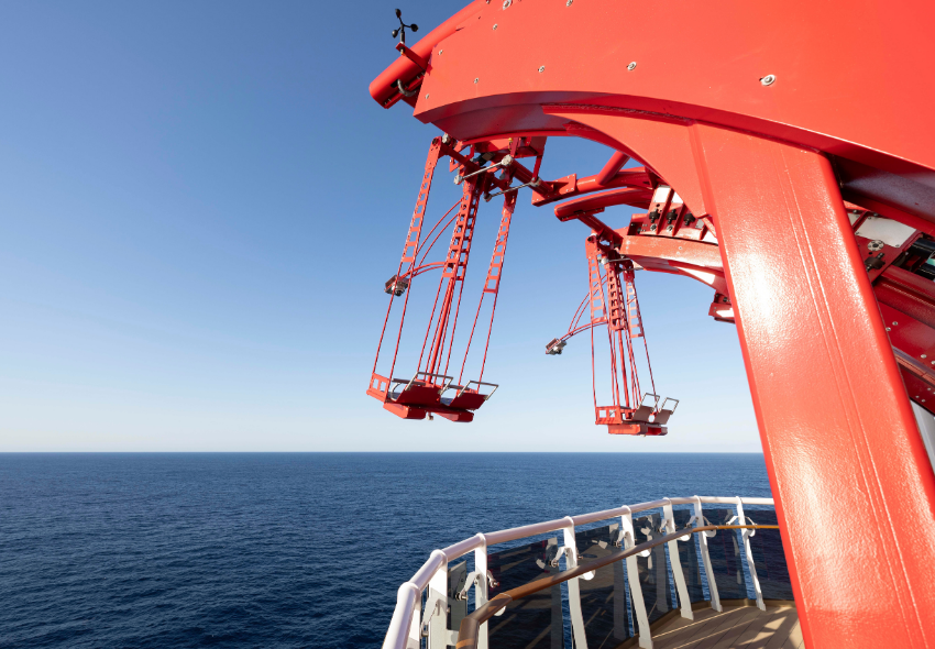 Auf dem Bild ist die Attraktion „Cliffhanger“ an Bord der MSC World Asia zu sehen. Mehrere rote Schaukelsitze sind an einer großen Konstruktion am Rand des Schiffes befestigt und ragen über die Reling hinaus. Im Hintergrund erstreckt sich das offene