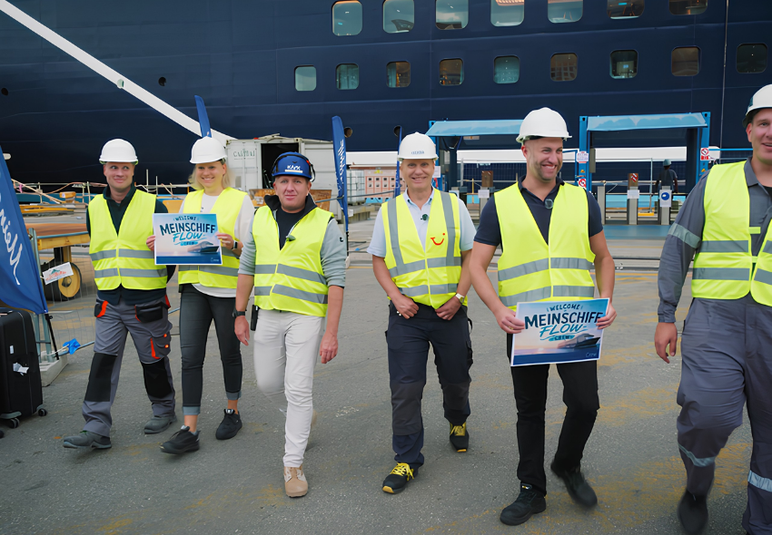 Mehrere Crew-Mitglieder der Mein Schiff Flow gehen über das Werftgelände und tragen weiße Schutzhelme sowie gelbe Warnwesten. Einige halten Willkommensschilder mit dem Logo der Mein Schiff Flow in den Händen. Im Hintergrund ist die dunkelblaue Außenw