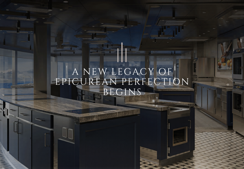 Das Bild zeigt die moderne Culinary Arts Kitchen an Bord der Seven Seas Prestige. Mehrere großzügige Kochstationen mit dunklen Schränken und hellen Arbeitsflächen sind in einem stilvoll gestalteten Raum angeordnet. Große Fenster lassen Tageslicht her