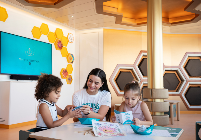 Kinder entdecken die Welt der Ozeane im Rahmen des Programms „Oceans Day powered by MSC Foundation“ Eine Mitarbeiterin der MSC Foundation sitzt an einem Tisch mit zwei Kindern in einem hellen, freundlich gestalteten Kinderbereich an Bord eines Kreuzfahrtschiffes. Gemeinsam beschäftigen sie sich mit Informationsmaterialien und Bastelunterlagen rund um d