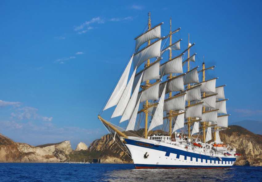 Star Clipper unter vollen Segeln im Mittelmeer Das Segelschiff Star Clipper fährt bei sonnigem Wetter mit vollständig gesetzten weißen Segeln über das tiefblaue Meer. Im Hintergrund erheben sich helle Felsformationen und sanfte Küstenhügel einer Mittelmeerinsel. Der weiße Rumpf mit blauen Akzen