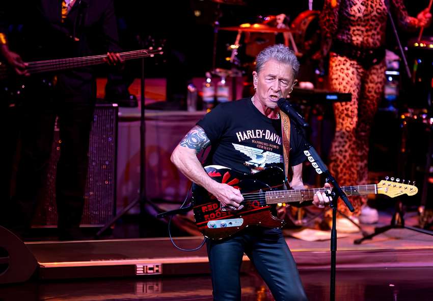 Peter Maffay begeistert Gäste bei Rock ’n’ Sail an Bord der Mein Schiff 1 Der Musiker Peter Maffay steht auf einer Konzertbühne an Bord der Mein Schiff 1 und singt in ein Mikrofon, während er E-Gitarre spielt. Die Bühne ist in kräftiges Rot- und Violettlicht getaucht. Im Hintergrund sind weitere Bandmitglieder und Instrumen