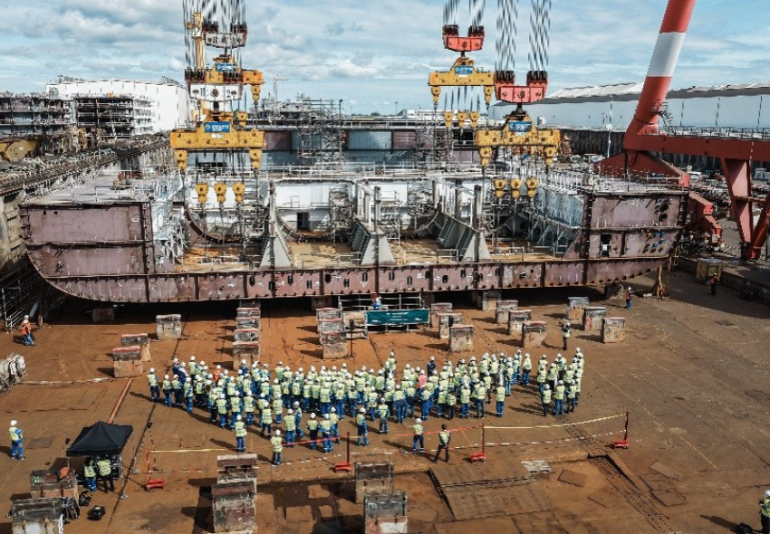 Kiellegung der Oasis 7 in Saint-Nazaire Die Aufnahme zeigt die traditionelle Kiellegungs-Zeremonie für die Oasis 7 in der Werft Chantiers de l’Atlantique im französischen Saint-Nazaire. Mehrere große Kräne halten einen massiven Stahlblock über dem künftigen Schiffsrumpf. Davor haben sic