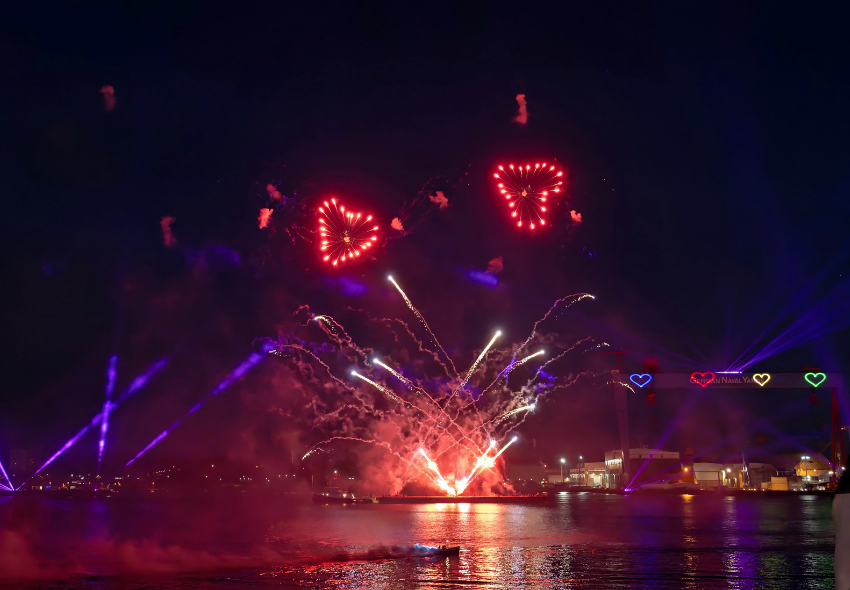 AIDA Abschlussinszenierung „Zwischen Himmel und Meer“ begeistert Besucher der Kieler Woche Bei Dunkelheit erhellen farbenfrohe Feuerwerks- und Lichteffekte den Himmel über dem Hafen. Über dem Wasser sind mehrere rote, herzförmige Feuerwerksmotive zu sehen, während sich helle Lichtstrahlen und Spiegelungen auf der Wasseroberfläche ausbreite