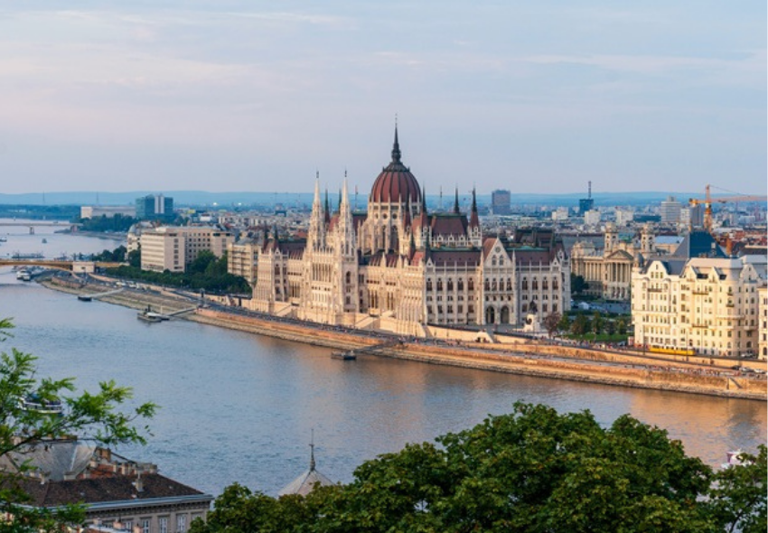 Budapest zählt zu den Höhepunkten ausgewählter Donaureisen mit nicko cruises Das Bild zeigt einen Blick auf das ungarische Parlamentsgebäude in Budapest direkt am Ufer der Donau. Das historische Gebäude mit seiner markanten Kuppel und zahlreichen Türmen erstreckt sich entlang des Flusses und wird von der Abendsonne angestrahlt.