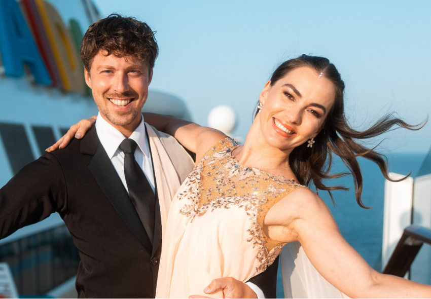 Renata und Valentin Lusin begleiten die Themenreise „AIDA tanzt“ 2027 Das Bild zeigt die Profitänzer Renata und Valentin Lusin, die gemeinsam an Deck eines Kreuzfahrtschiffes posieren. Renata Lusin trägt ein helles, mit funkelnden Verzierungen besetztes Kleid und lächelt in die Kamera. Valentin Lusin steht neben ihr in e