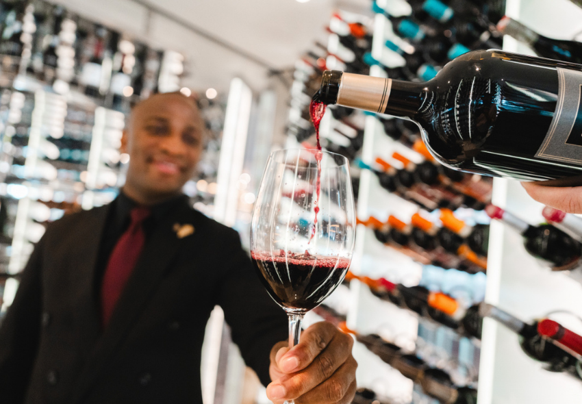 Sommelier präsentiert Rotwein an Bord von Celebrity Cruises Ein Sommelier hält ein Weinglas, während Rotwein aus einer Flasche eingeschenkt wird. Im Hintergrund sind zahlreiche Weinflaschen in einem modernen Weinraum zu sehen. Das Bild steht für das mehrfach ausgezeichnete Weinprogramm von Celebrity Cruises und