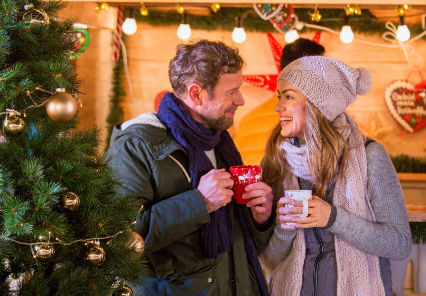Ein Paar steht auf einem festlich geschmückten Weihnachtsmarkt und hält warme Getränke in den Händen. Im Vordergrund ist ein dekorierter Weihnachtsbaum mit goldenen Kugeln zu sehen, während im Hintergrund Lichterketten und weihnachtliche Dekorationen