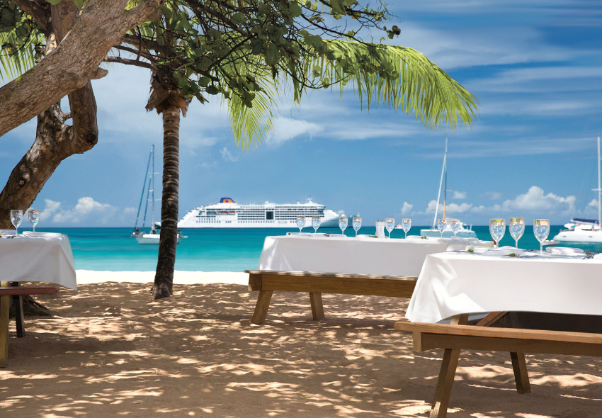 Exotische Adventszeit mit der EUROPA 2 und HANSEATIC spirit Im Hintergrund ein Schiff von Hapag-Lloyd Cruises, welches in der Karibik im Meer liegt, im Vordergrund Palmen und eingedeckte Tische mit weißen Decken am Strand