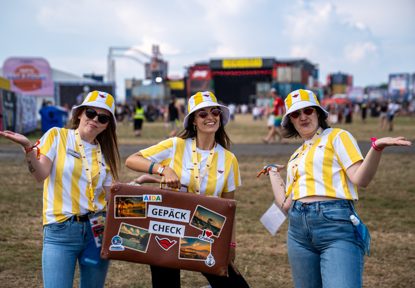 Drei Frauen im gelb weiß gestreiften Shirts und Hut halten den AIDA Gepäck Check Koffer in die Kamera vor dem Hintergrund einer Festival Bühne