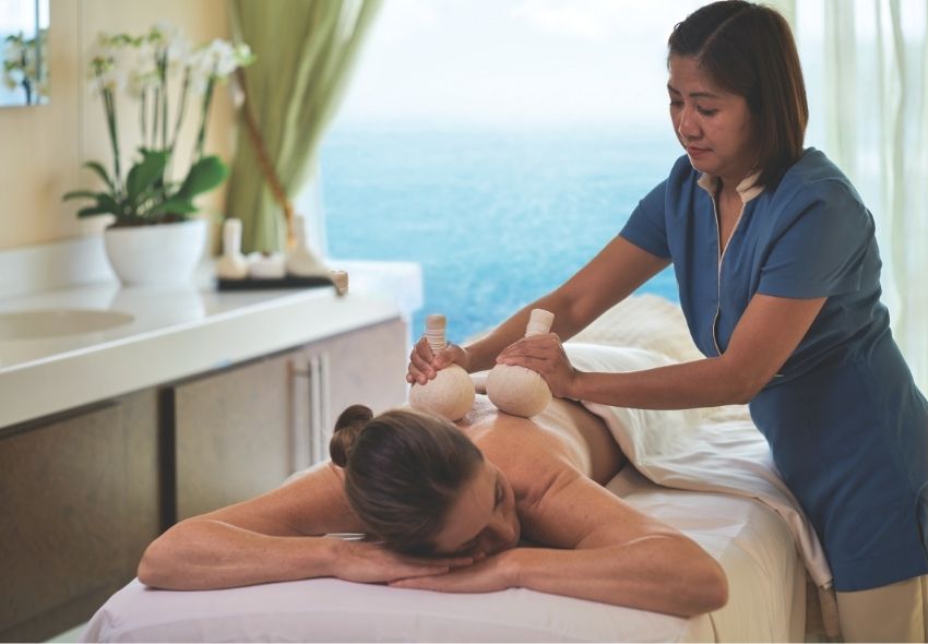 Wellness-Anwendung an Bord von Oceania Cruises Eine Spa-Mitarbeiterin massiert den Rücken einer entspannt auf einer Liege liegenden Frau mit warmen Kräuterstempeln. Durch das Fenster ist das Meer zu sehen. Der helle Behandlungsraum mit Orchideen und sanften Farben vermittelt eine ruhige und erholsam