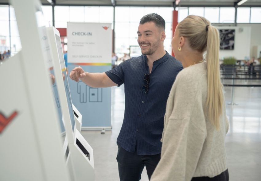 Digitaler Self-Service-Check-in im Kreuzfahrtterminal Ein Mann und eine Frau stehen vor einem digitalen Check-in-Terminal im Cruise Terminal und bedienen gemeinsam den Touchscreen. Im Hintergrund weist ein Schild auf den Check-in-Bereich hin. Der digitale Self-Service ermöglicht Gästen eine schnelle und ei