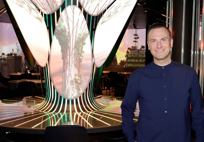 Sternekoch Tim Raue steht lächelnd im Restaurant FUGU an Bord der Mein Schiff Flow. Hinter ihm ist die auffällige digitale Installation im Stil eines stilisierten Baumes zu sehen, die das Restaurantkonzept mit Licht- und Projektionseffekten prägt und d