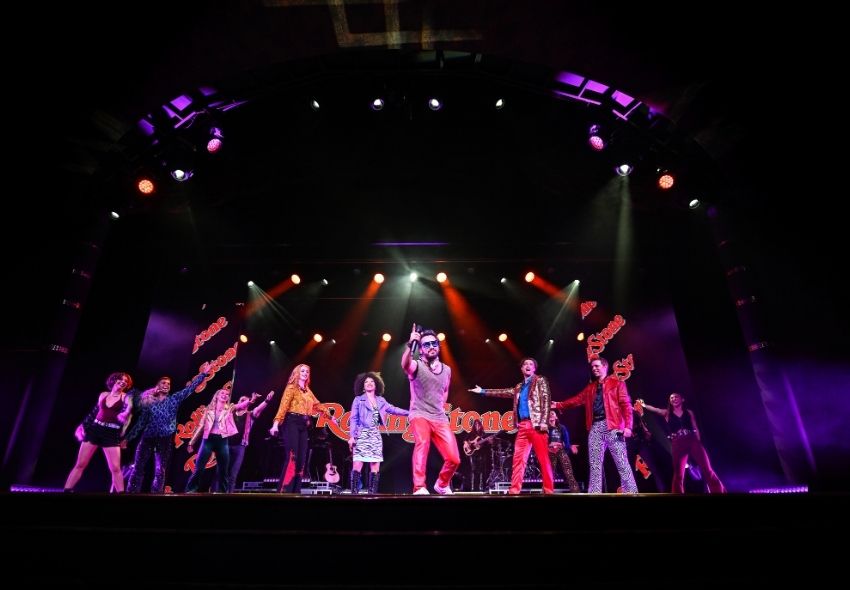 Mehrere Sänger und Tänzer stehen während einer Bühnenshow auf der World Stage der Oosterdam. Farbige Scheinwerfer tauchen die Bühne in rotes und violettes Licht, während im Hintergrund das Logo von Rolling Stone zu sehen ist. Die aufwendig inszenier