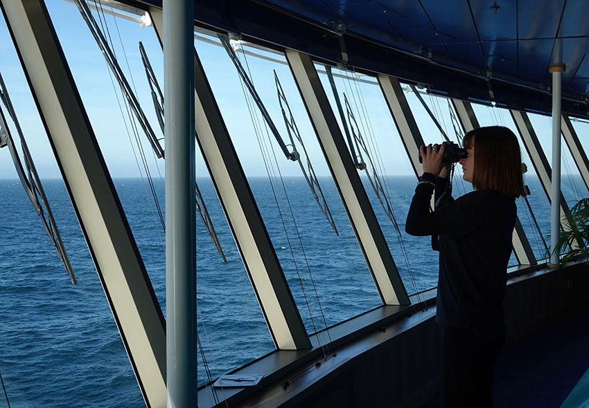 Maria Snell schaut mit Fernglas auf der Brücke aufs Meer