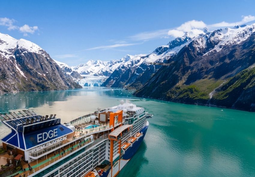 Celebrity Edge vor der beeindruckenden Gletscherkulisse Alaskas Die Celebrity Edge fährt durch eine weitläufige Bucht in Alaska. Das Kreuzfahrtschiff ist von türkisfarbenem Wasser und steilen, teilweise schneebedeckten Bergen umgeben. Im Hintergrund erstreckt sich ein Gletscher zwischen den Bergmassiven und unterst