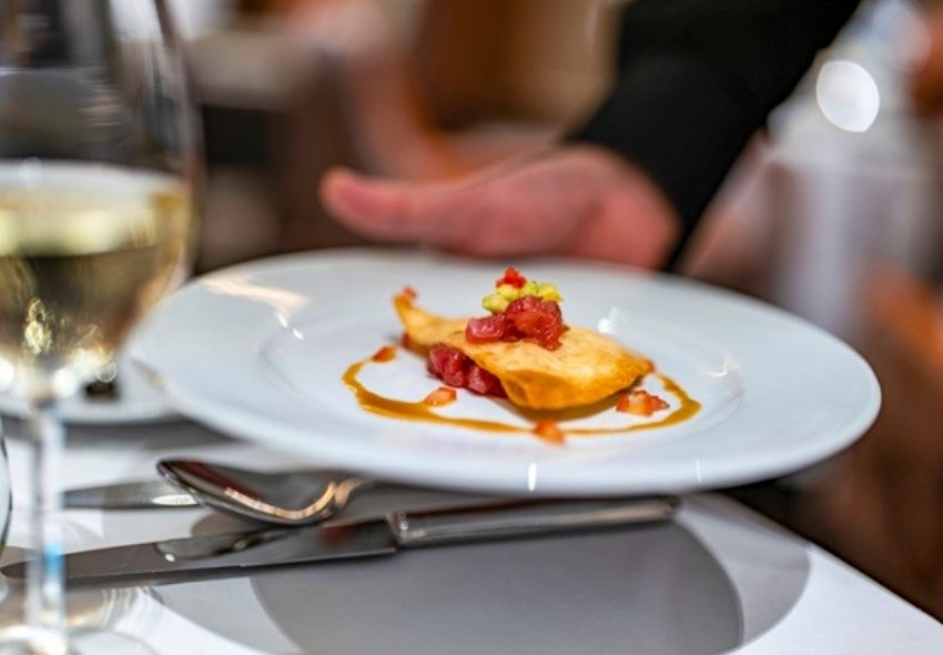Genuss auf höchstem Niveau an Bord der nickoSPIRIT Auf einem gedeckten Tisch steht ein weißer Teller mit einem angerichteten Gourmetgericht. Im Hintergrund ist eine Person zu sehen, daneben steht ein Glas Weißwein.