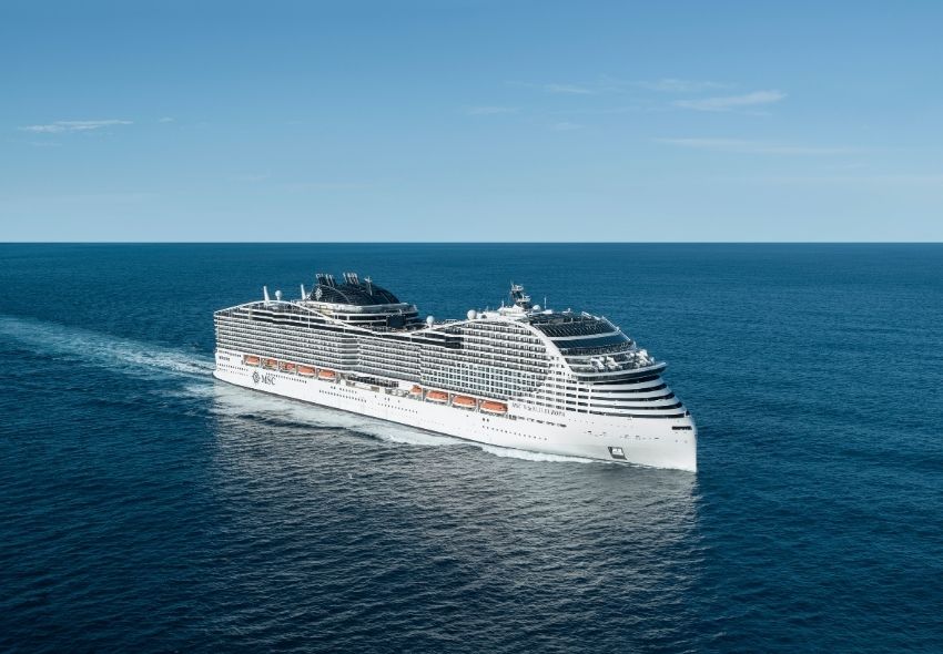 Die MSC World Europa fährt bei klarem Himmel über das tiefblaue Meer. Das große weiße Kreuzfahrtschiff ist aus erhöhter Perspektive schräg von vorne zu sehen.