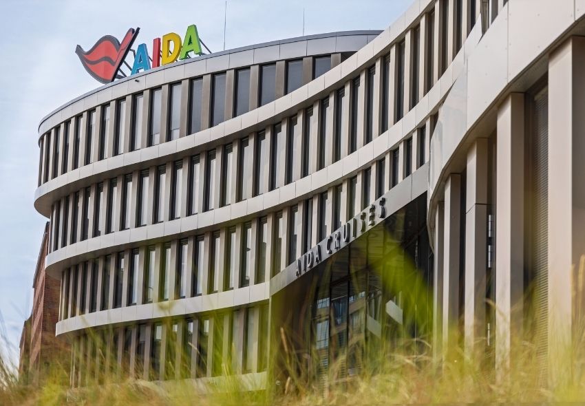 AIDA Headquarter in Rostock Das moderne AIDA Headquarter in Rostock mit geschwungener Gebäudefassade. Auf dem Dach ist das farbige AIDA Logo zu sehen, über dem Eingangsbereich der Schriftzug „AIDA Cruises“.