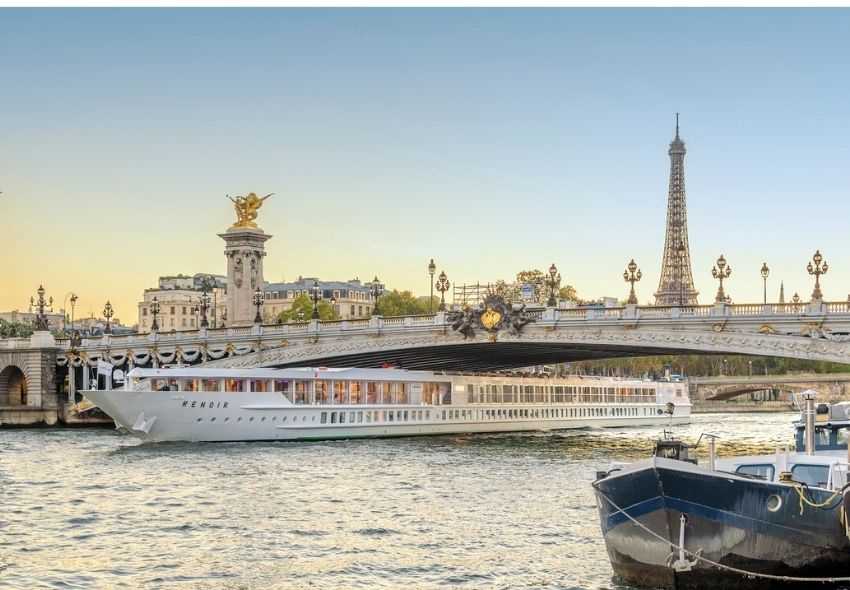 Die MS Renoir von CroisiEurope fährt auf der Seine in Paris unter der Pont Alexandre III hindurch. Im Hintergrund ist der Eiffelturm zu sehen, im Vordergrund liegen weitere Boote am Ufer.