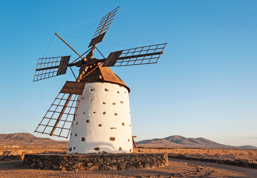 Traditionelle Windmühle auf Fuerteventura Eine traditionelle weiß getünchte Windmühle mit dunklen Holzflügeln steht in der kargen Landschaft Fuerteventuras. Im Hintergrund erstrecken sich sanfte, braune Vulkanberge unter einem klaren blauen Himmel.
