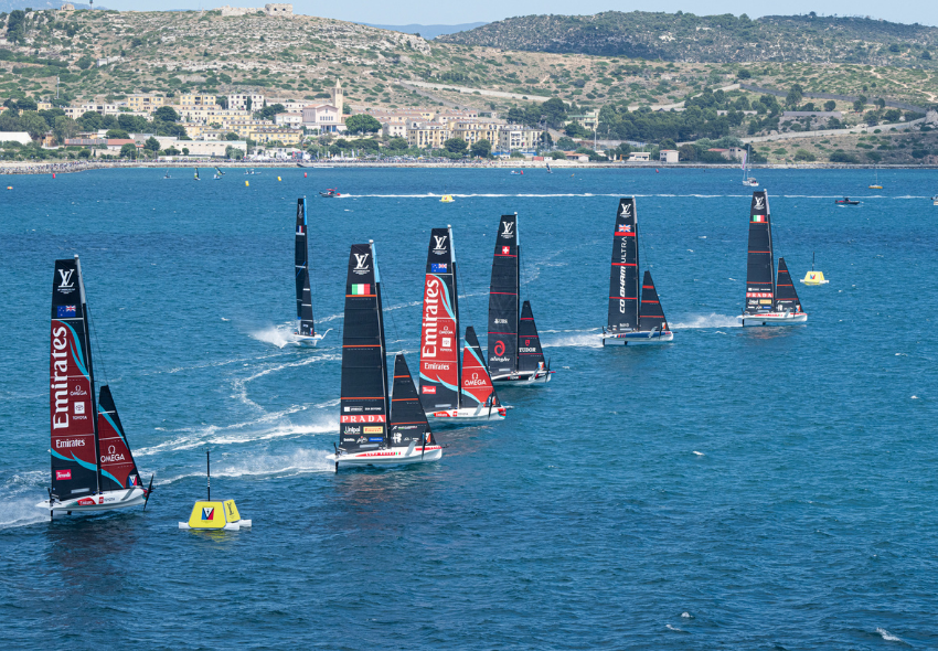 America’s Cup – Segelboote während einer Regatta Mehrere schwarze Hochleistungs-Segelboote fahren während einer America’s-Cup-Regatta über das blaue Meer. Die Boote sind hintereinander auf dem Wasser verteilt und ziehen weiße Gischtspuren hinter sich her. Im Hintergrund ist eine hügelige Küstenla