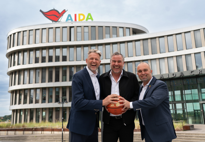 AIDA Cruises und ROSTOCK SEAWOLVES setzen Partnerschaft fort Drei lachende Männer stehen mit einem Basketball vor dem AIDA Headquarter in Rostock. Gemeinsam halten sie den Ball in die Kamera, während über ihnen das große AIDA Logo am Gebäude zu sehen ist.