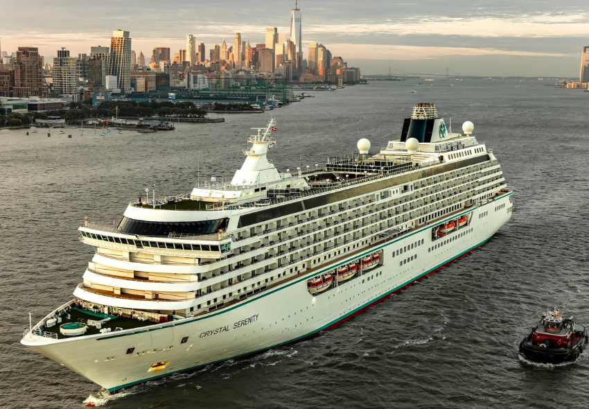 Außenansicht der Crystal Serenity auf dem Wasser mit der Skyline von New York links im Hintergrund, rechts ein Pilotboot