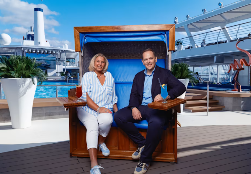 Tanya Kumst und Denis Wiechert präsentieren neuen Mein Schiff Kulinarik-Podcast Eine blonde Frau in einer hellblau-weiß gestreiften Bluse und ein Mann in einem dunkelblauen Anzug sitzen lächelnd nebeneinander in einem großen Strandkorb mit blauen Polstern. Im Hintergrund sind ein Swimmingpool, Palmen in weißen Pflanzkübeln und d