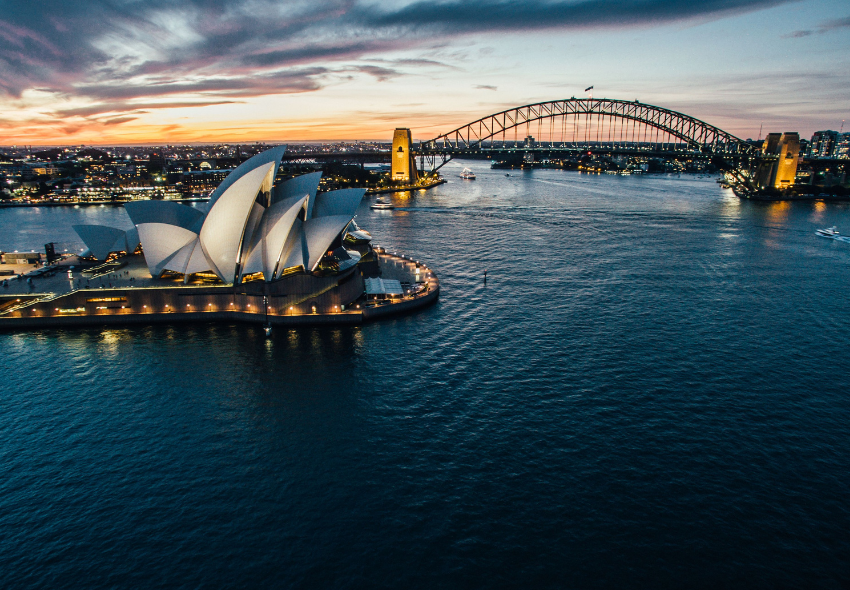 Luftaufnahme des Hafens von Sydney in der Abenddämmerung. Im Vordergrund liegt das beleuchtete Sydney Opera House mit seinen markanten, segelförmigen Dächern. Dahinter überspannt die Sydney Harbour Bridge das Wasser. Am Horizont leuchtet der Himmel in