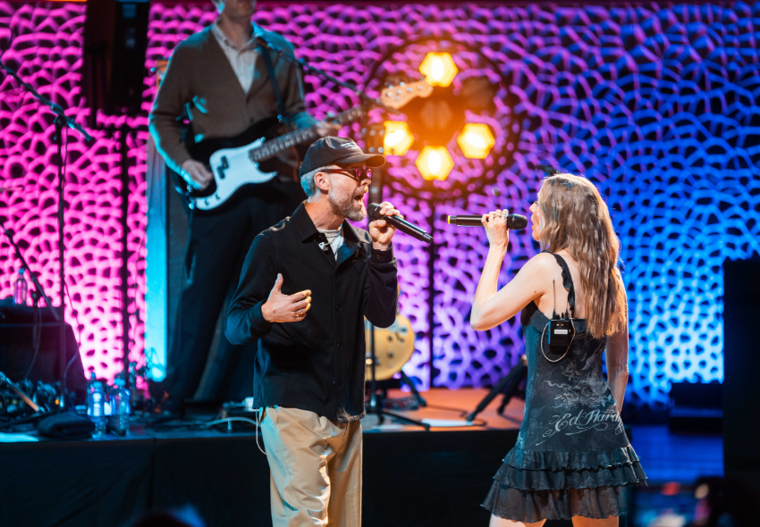 Mark Forster und LEA beim AIDA & Friends Konzert in der Elbphilharmonie Ein männlicher Sänger mit schwarzer Kappe, Brille und dunkler Jacke und eine Sängerin mit langen braunen Haaren und schwarzem Kleid stehen sich auf der Bühne gegenüber und singen in ihre Mikrofone. Im Hintergrund spielt ein Musiker E-Bass. Die Bühne